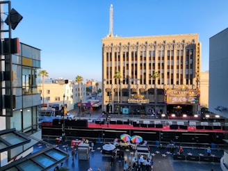 A bustling urban scene featuring a historic theater building with an ornate facade and a marquee lit up with bright lights. In the foreground, a bustling area with people walking on a star-lined sidewalk, possibly representing a famous tourist destination. There are vendors with colorful umbrellas and a barrier sectioned off area suggesting preparations for an event. The sky is clear and blue, adding to the vibrant atmosphere.