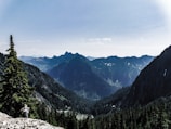 A scenic view of a densely forested valley surrounded by towering mountain peaks under a clear blue sky. A hiker is visible in the foreground, traversing rocky terrain with a backpack, suggesting a sense of adventure and exploration.