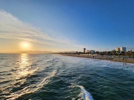 A beautiful coastline with gentle waves meeting the sandy beach, lined with numerous people enjoying leisure activities. The sun is setting on the horizon, casting a warm golden glow over the water and silhouetting a distant range of mountains. Several high-rise buildings are visible along the shoreline, further adding to the vibrant atmosphere of the scene.