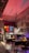 A warmly lit, eclectic cafe interior with vintage decor. The concrete walls are adorned with various framed art pieces and illuminated by a red lamp. There are tables and chairs, and a counter with various items and a refrigerator. The atmosphere is cozy with a mix of modern and rustic elements.