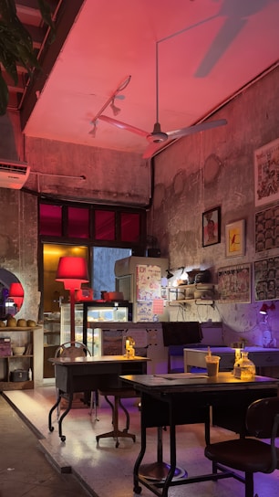 A warmly lit, eclectic cafe interior with vintage decor. The concrete walls are adorned with various framed art pieces and illuminated by a red lamp. There are tables and chairs, and a counter with various items and a refrigerator. The atmosphere is cozy with a mix of modern and rustic elements.
