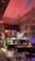 A warmly lit, eclectic cafe interior with vintage decor. The concrete walls are adorned with various framed art pieces and illuminated by a red lamp. There are tables and chairs, and a counter with various items and a refrigerator. The atmosphere is cozy with a mix of modern and rustic elements.