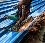 A person is using an angle grinder to cut metal roofing sheets. The device emits bright, orange sparks that contrast with the blue of the sheets. The person is wearing a protective glove on one hand and has a protective covering on one of their legs.