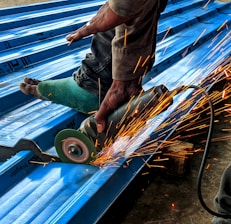 A person is using an angle grinder to cut metal roofing sheets. The device emits bright, orange sparks that contrast with the blue of the sheets. The person is wearing a protective glove on one hand and has a protective covering on one of their legs.