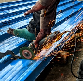 A person is using an angle grinder to cut metal roofing sheets. The device emits bright, orange sparks that contrast with the blue of the sheets. The person is wearing a protective glove on one hand and has a protective covering on one of their legs.