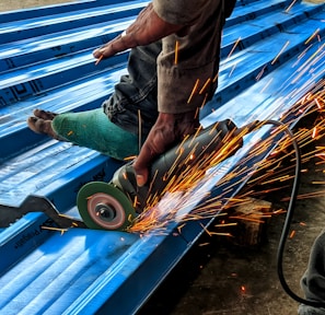 A person is using an angle grinder to cut metal roofing sheets. The device emits bright, orange sparks that contrast with the blue of the sheets. The person is wearing a protective glove on one hand and has a protective covering on one of their legs.