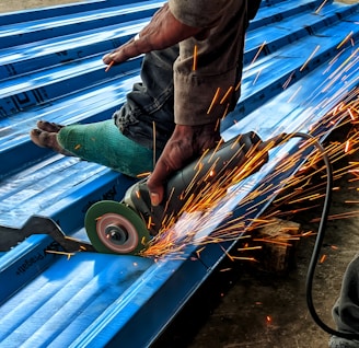 A person is using an angle grinder to cut metal roofing sheets. The device emits bright, orange sparks that contrast with the blue of the sheets. The person is wearing a protective glove on one hand and has a protective covering on one of their legs.