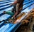 A person is using an angle grinder to cut metal roofing sheets. The device emits bright, orange sparks that contrast with the blue of the sheets. The person is wearing a protective glove on one hand and has a protective covering on one of their legs.