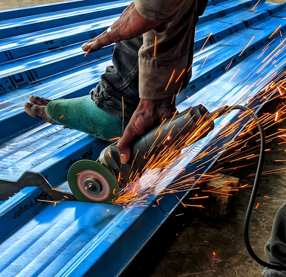 A person is using an angle grinder to cut metal roofing sheets. The device emits bright, orange sparks that contrast with the blue of the sheets. The person is wearing a protective glove on one hand and has a protective covering on one of their legs.