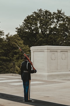 A member of the military stands in full uniform, holding a rifle upright at a ceremonious location, presumably a memorial or sacred site. The soldier is positioned beside a white marble structure with inscribed text, surrounded by lush green trees.
