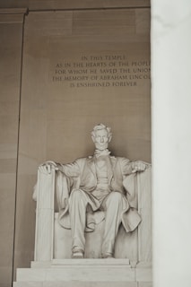 A large marble statue of Abraham Lincoln seated on a grand chair within a monumental setting. The inscription above the statue honors Lincoln's memory as a savior of the Union, enshrined forever. The environment exudes grandeur and reverence.
