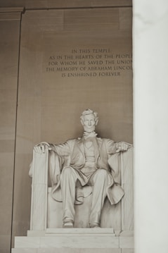 A large marble statue of Abraham Lincoln seated on a grand chair within a monumental setting. The inscription above the statue honors Lincoln's memory as a savior of the Union, enshrined forever. The environment exudes grandeur and reverence.