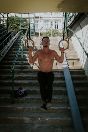 A shirtless man exercises on gymnastic rings in an outdoor urban setting, positioned on a staircase with metal railings and a bag nearby. The background features buildings and greenery.