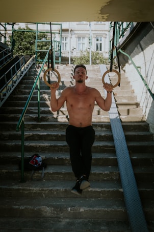 A shirtless man exercises on gymnastic rings in an outdoor urban setting, positioned on a staircase with metal railings and a bag nearby. The background features buildings and greenery.