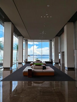 A spacious, modern lobby area featuring large floor-to-ceiling windows that overlook a scenic view of the ocean and distant islands. The interior includes several contemporary lounge sofas arranged symmetrically, set on a glossy floor that reflects the natural light. The walls are lined with vertical pillars, adding to the grandeur and openness of the space.