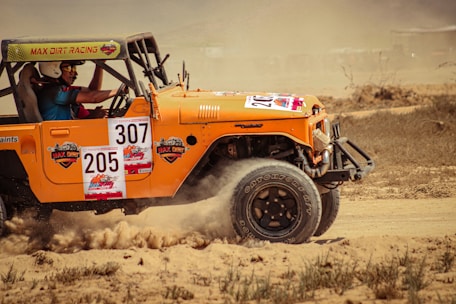 An orange off-road vehicle is racing through a dusty terrain, kicking up dirt as it moves. The driver is focused, wearing a helmet and a blue shirt. Several racing stickers and numbers are displayed on the vehicle, indicating its participation in a competitive event.