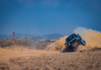 A rugged off-road vehicle is kicking up a large cloud of dust as it speeds through a desert terrain with a dramatic incline. The blue sky and distant mountains provide a scenic backdrop, while a red flag on a stick can be seen to the left.