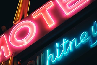 Bright neon signs featuring the word 'MOTEL' in vibrant pink and yellow, along with another word that appears to be 'Whitney' in blue. The signs emit a colorful glow against a dark background, creating a retro and vibrant atmosphere.
