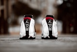 A pair of white athletic shoes with black and red accents, featuring a prominent number 23 and a logo on the heel. The shoes are positioned on a textured surface with an urban background out of focus.