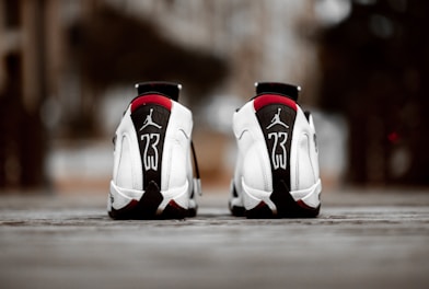 A pair of white athletic shoes with black and red accents, featuring a prominent number 23 and a logo on the heel. The shoes are positioned on a textured surface with an urban background out of focus.
