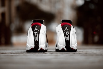 A pair of white athletic shoes with black and red accents, featuring a prominent number 23 and a logo on the heel. The shoes are positioned on a textured surface with an urban background out of focus.
