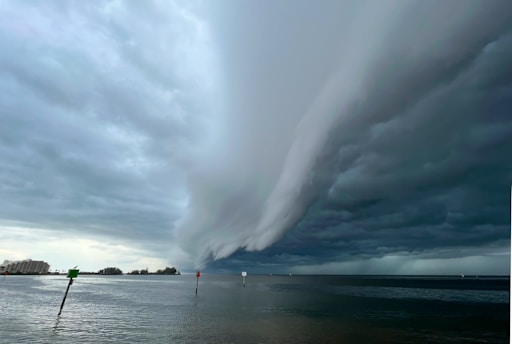 Dark clouds forming a massive, ominous storm front over a body of water with some navigational markers visible. The storm clouds are dramatic, taking up most of the sky, with a slight bit of light breaking through on the left side near the horizon. On the left edge, some buildings are also visible.