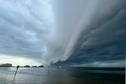 Dark clouds forming a massive, ominous storm front over a body of water with some navigational markers visible. The storm clouds are dramatic, taking up most of the sky, with a slight bit of light breaking through on the left side near the horizon. On the left edge, some buildings are also visible.