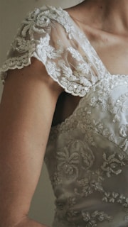 Close-up of a person wearing an intricately embroidered, lace-covered garment. The lace is detailed with floral patterns and covers the shoulder and upper arm area, featuring a delicate, feminine design.