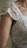 Close-up of a person wearing an intricately embroidered, lace-covered garment. The lace is detailed with floral patterns and covers the shoulder and upper arm area, featuring a delicate, feminine design.