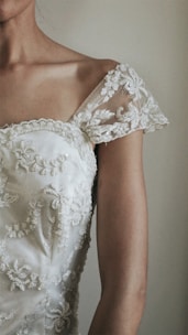 A close-up of a woman's shoulder wearing a delicate white lace dress. The fabric is embroidered with intricate floral patterns and features a sheer lace cap sleeve. The image captures a minimalist and elegant aesthetic.