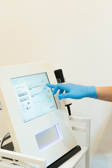 A hand wearing a blue glove is interacting with a touchscreen monitor. The device appears to be part of medical or laboratory equipment. The interface displays various settings and options.
