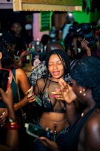 A lively party scene with individuals gathered closely together. People are holding smartphones and drinks, capturing moments and engaging with each other. The environment is vibrant with a dimly lit, colorful background and a dense crowd.