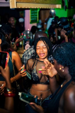 A lively party scene with individuals gathered closely together. People are holding smartphones and drinks, capturing moments and engaging with each other. The environment is vibrant with a dimly lit, colorful background and a dense crowd.