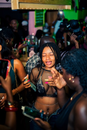 A lively party scene with individuals gathered closely together. People are holding smartphones and drinks, capturing moments and engaging with each other. The environment is vibrant with a dimly lit, colorful background and a dense crowd.