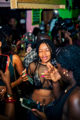 A lively party scene with individuals gathered closely together. People are holding smartphones and drinks, capturing moments and engaging with each other. The environment is vibrant with a dimly lit, colorful background and a dense crowd.