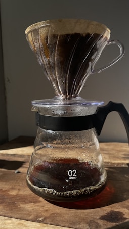 A pour-over coffee setup with a glass dripper sitting atop a glass carafe. The coffee dripper contains dark brown coffee grounds and a paper filter, with coffee dripping into the carafe below. The setup is placed on a wooden surface and is illuminated by natural sunlight, casting soft shadows.