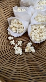 A close-up view of several paper bags filled with popcorn arranged in a woven basket. There are also some popcorn kernels scattered in the basket.