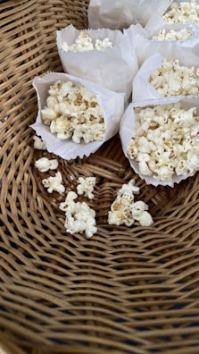A close-up view of several paper bags filled with popcorn arranged in a woven basket. There are also some popcorn kernels scattered in the basket.