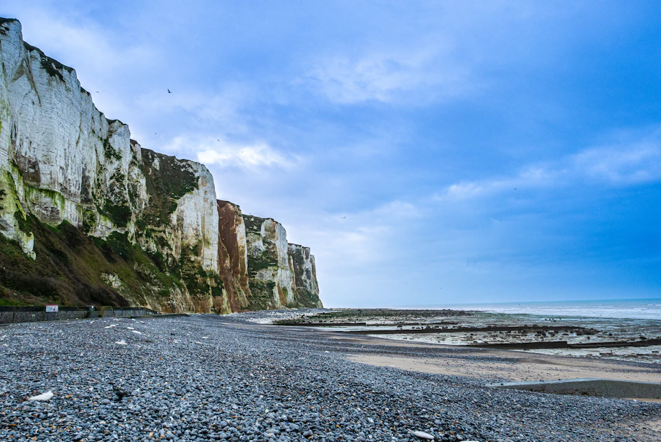 The image depicts a rocky beach with white cliffs on either side, creating a picturesque coastal scene. The cliffs are covered in green vegetation and have a reddish-brown hue, adding depth to the landscape. The beach is sandy, contrasting with the ruggedness of the cliffs. In the distance, beyond the cliffs, there's an ocean visible at the horizon line.