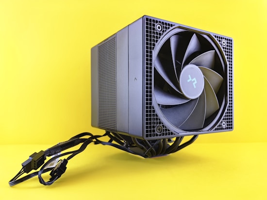 A computer cooling fan or air cooler with a large, black circular fan is connected with multiple cables and placed against a bright yellow background. The design is sleek and modern, featuring a metallic grid structure.