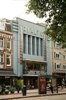 A classic cinema building with the name 'RIALTO' displayed at the top. The structure features an art deco style with vertical lines and a teal facade, accompanied by brown brick buildings on either side. There is a café or restaurant on the ground level with a few people outside, one on a scooter. A large poster and a public transit stop are visible in the foreground. Trees partially obscure the view of the roofline.