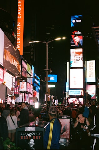 A bustling nighttime scene in a busy city area filled with bright lights and billboards. Crowds of people walk through the streets, surrounded by towering buildings and vibrant advertisements. The atmosphere is lively and energetic, capturing the essence of an urban nightlife.