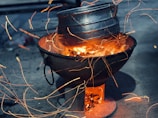 A metal pot rests on top of a traditional open charcoal or wood stove. Bright orange sparks and flames are visible, indicating an active fire. The scene is set on a gray concrete surface, creating a warm and dynamic contrast between the fire and the cool surroundings.