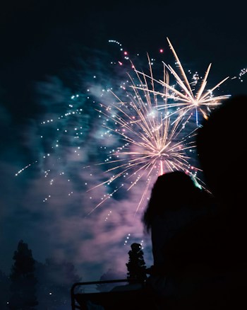 A vibrant display of fireworks illuminates the night sky, with colorful bursts and sparkles creating an energetic spectacle. Silhouettes of people and trees are visible in the foreground, adding a sense of scale and anticipation.