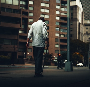 In an urban setting, a person with a cane walks on a sidewalk near tall, residential buildings. The atmosphere appears dim, with a hint of isolation and solitude.