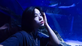 A person gazes thoughtfully upward in an aquarium setting, with various shades of blue lighting creating an ethereal atmosphere. The reflective surfaces and aquatic environment surrounding them contribute to a contemplative mood.