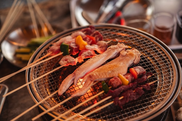 A barbecue grill with various skewered meats and vegetables cooking over a flame. The skewers consist of chicken wings, beef, and colorful peppers. The background shows blurred kitchen utensils and ingredients, hinting at a meal preparation setting.