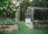 A greenhouse constructed with glass panes and a stone base is located on a green lawn. To the left, there are garden beds surrounded by wooden fences, filled with a variety of plants and vines carefully tended with support structures. Tall trees with dense foliage create a lush background, giving the area a sense of natural tranquility.