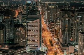 A bustling cityscape at night with tall skyscrapers lining a lit street that stretches into the distance. The buildings glow with numerous lights, reflecting the vibrant urban atmosphere.