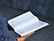 A person holding an open book with Arabic script on its pages. The book appears to be religious in nature, possibly the Quran. The person is wearing a dark blue garment with visible sleeves.